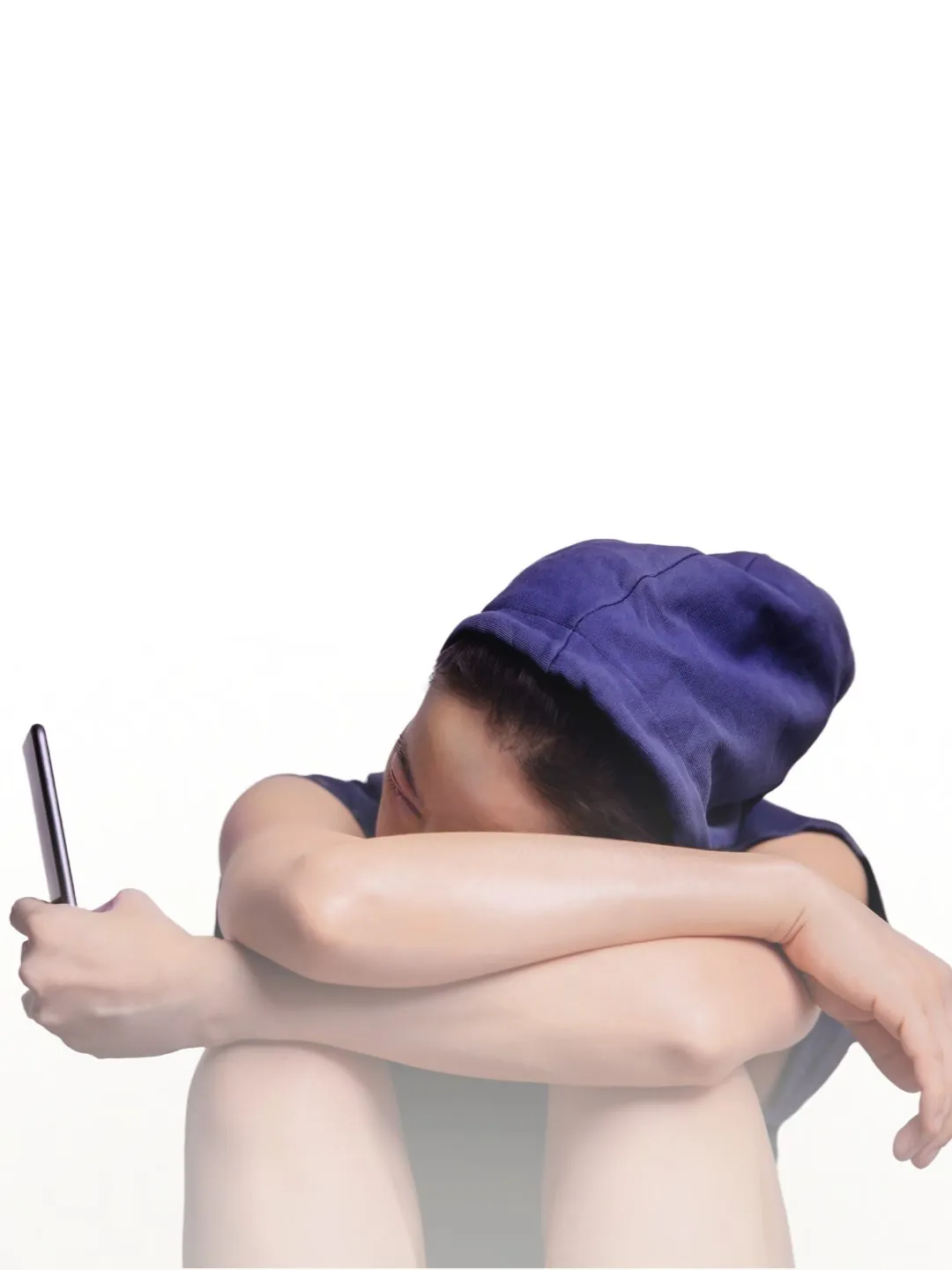 Jovem sentado com a cabeça apoiada entre os braços e as pernas, segurando um smartphone, em postura de isolamento e tristeza.
